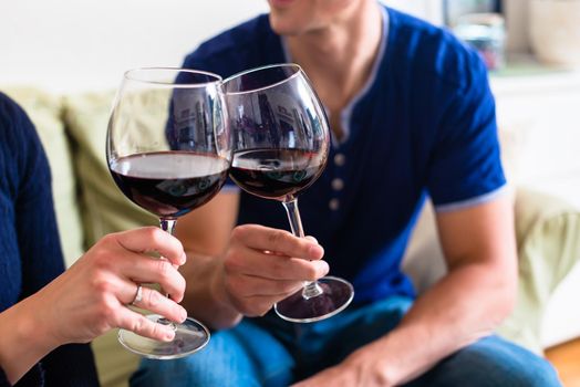 Romantic young couple toasting with red wine in stemmed glasses while sitting on the couch at home