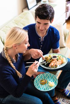 Young Caucasian couple eating Asian traditional food with chopsticks while sitting on the couch at home