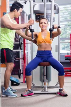 Instructor supervising sportive woman during machine workout at butterfly station in fitness center