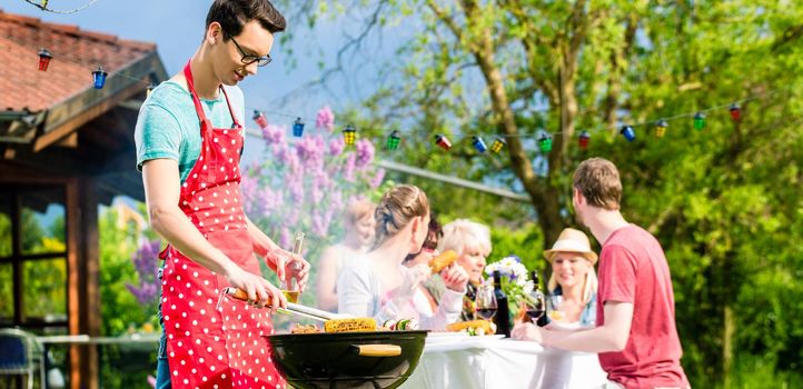 Man grilling meat on garden barbecue party, in the background friends eating and drinking