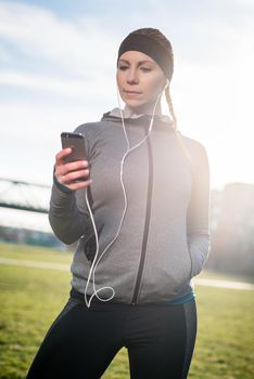 Beautiful thoughtful young woman listening to music on her mobile phone while wearing trendy sport outfits in a sunny day in the park