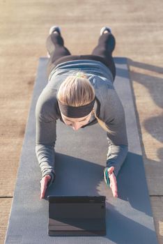 High-angle view of a fit young woman watching a motivational video on tablet PC, while exercising the forearm plank position on a mat outdoors in the park