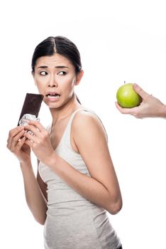 Funny young woman eating a fresh apple while looking at a chocolate bar