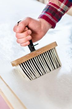 Close-up of the hand of a man holding a synthetic brush with plastic handle while applying paste to the surface of the wallpaper sheet