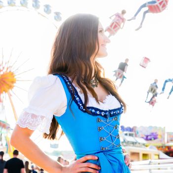 Dirndl wearing woman is standing in front of ferris wheel and carousel at Bavarian folk festival