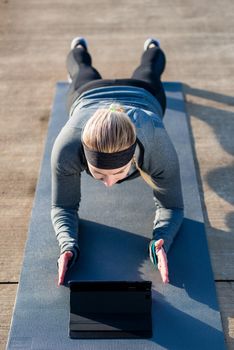 High-angle view of a fit young woman, watching a motivational video on tablet PC while exercising the forearm plank position on a mat outdoors in the park
