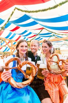 Three friends in beer tent at Dult or Oktoberfest holding giant pretzels up in the air