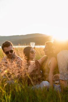 Friends enjoying evening mood of summer day sitting in high grass with drinks and guitar music