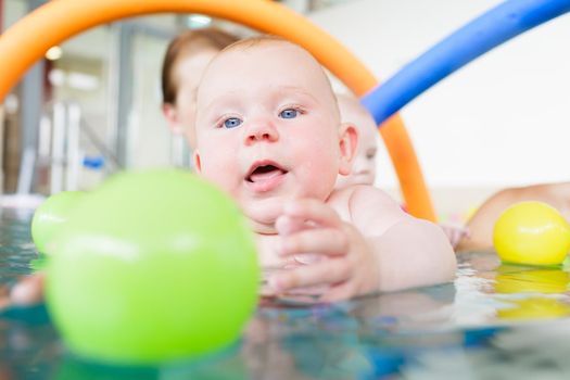 Newborn baby in pool at infant swimming lesson reaching for water ball