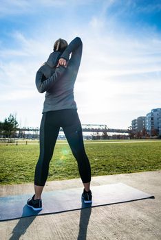 Rear view of a fit woman practicing a shoulder stretch as routine exercise for warming up before outdoor workout in the park