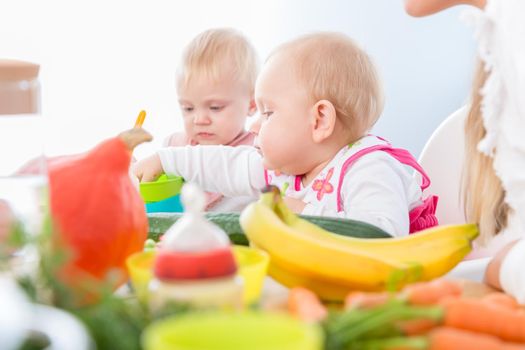 Portrait of a cute baby girl with blue eyes eating healthy solid food, while sitting in a high chair next to another baby in a modern daycare center for babies