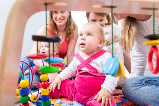 Portrait of a cute baby girl looking up with her beautiful blue eyes, while sitting down on the floor with other two babies and their mothers at home