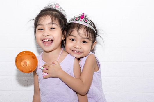 Female asian identical twins sitting on chair with white background. Wearing purple dress and accessories. Hugging each other while standing and smiling