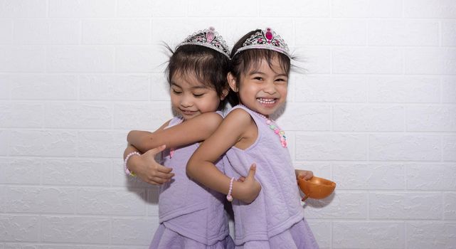 Female asian identical twins sitting on chair with white background. Wearing purple dress and accessories. Standing back to back with each other and having fun