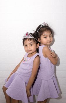 Female asian identical twins sitting on chair with white background. Wearing purple dress and accessories. Standing back to back with each other and having fun