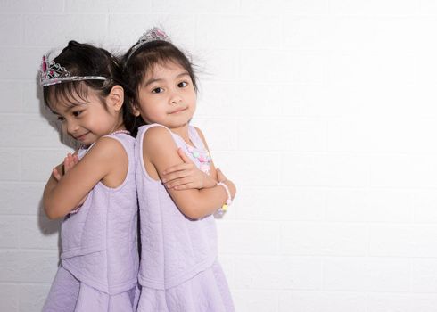 Female asian identical twins sitting on chair with white background. Wearing purple dress and accessories. Standing back to back with each other and having fun