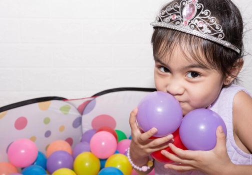 Female asian child girl  while sitting and playing with colorful plastic balls while wearing accessories like crown and necklace