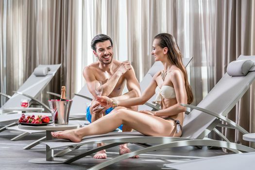 Romantic young man opening a bottle of champagne while relaxing with his partner at the swimming pool in a fancy hotel