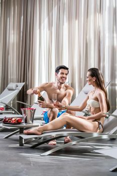 Romantic young man opening a bottle of champagne while relaxing with his partner at the swimming pool in a fancy hotel