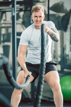 Full length low angle view of a handsome bodybuilder exercising with heavy battle ropes during intense functional training workout at the gym