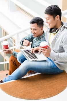Two young Asian men using a laptop while relaxing during break on the stairs of a modern building