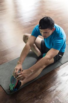 Low-angle full length view of a man sitting down on exercise mat while touching his toes during stretching routine at the gym