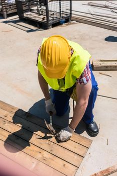 High-angle view of a blue-collar worker wearing yellow hard hat, safety vest and gloves while using a hammer during work on the construction site