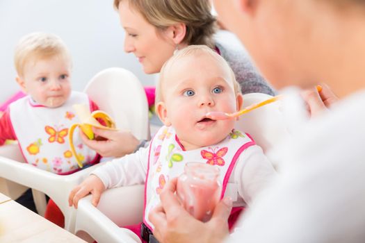 Portrait of a cute and healthy baby girl with blue eyes looking at her mother, while eating nutritious fruit puree next to another baby at home