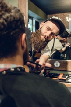 Close-up side view of the head of a young man and the hands of a skilled hairstylist trimming his beard, with comb and scissors in a cool hair salon