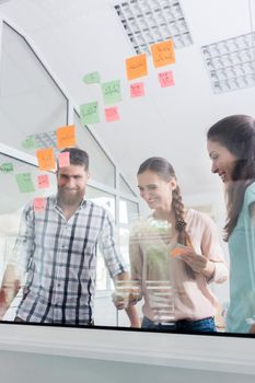 Low-angle view of reliable young workers posting reminders on the transparent wall of an office, next to their colleague in a modern co-working space