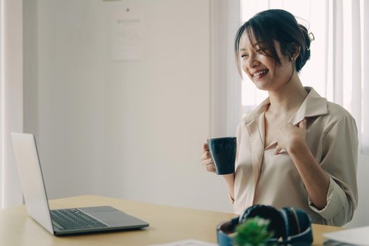 Attractive asian woman having video call via laptop while hand hold a cup of coffee at home office, Consultation, webinar, tutoring on internet, telecommuting concept