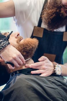 Close-up of the hand of a skilled barber using scissors, while trimming the beard of a young redhead customer in a trendy hair salon for men
