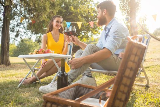 Young and beautiful couple in love toasting with red wine while sitting down on folding chairs during romantic picnic in the countryside