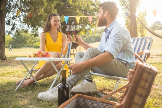 Young and beautiful couple in love toasting with red wine while sitting down on folding chairs during romantic picnic in the countryside