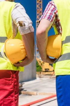 Close-up rear view of the hands of two workers wearing reflective safety vests while holding yellow hard hats outdoors on the construction site