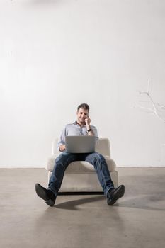 Young man sitting in empty room, using cell phone and notebook PC