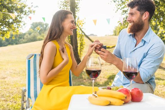 Beautiful young woman accepting with joy and emotion the marriage proposal from her boyfriend during romantic picnic in a sunny day of summer