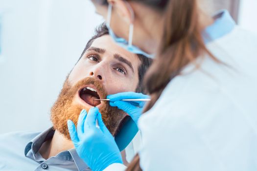 Side view of a reliable female dentist, using sterile instruments and blue surgical gloves while cleaning the teeth of a patient in the dental office of a modern clinic