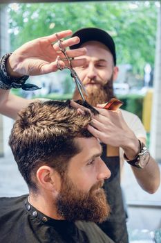 Dedicated male hairstylist using scissors and plastic comb while giving a cool haircut to a redhead bearded young man in a trendy beauty salon