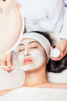 Close-up of the face of a young woman, relaxing under the gentle touch of the specialist applying on her cheeks white facial mask with rejuvenating effects