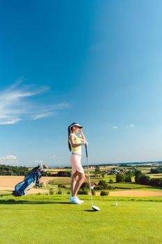 Side view of a fit woman wearing modern golf outfits, while looking at the horizon on the green grass of a golf course in the countryside in a sunny day of summer