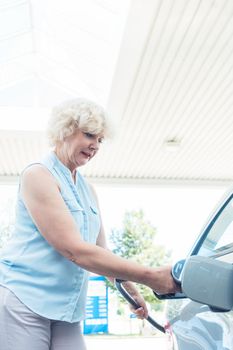 Low-angle view portrait of an active senior woman smiling while filling up the gas tank of her car at the station in summer