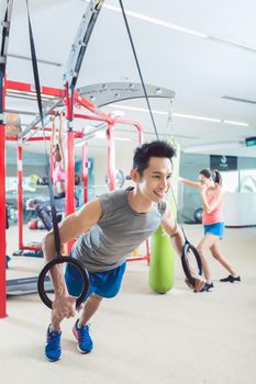 Cheerful fit young man exercising with gymnastic rings during routine workout in a trendy fitness center with modern equipment for functional training