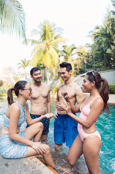 Multi-ethnic group of four young and cheerful friends smiling and talking while relaxing together at a trendy outdoor swimming pool in summer