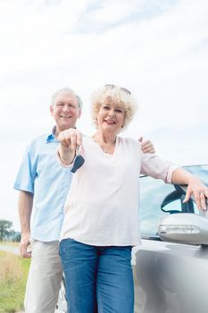 Close-up of the hands of a senior woman showing the keys of her car while posing next to her partner as confident elderly drivers