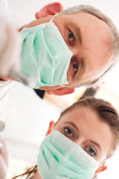 Dental treatment with dentist and dental assistant bowing over the patient as seen from patient's perspective. They have drills and angled mirrors, focus on eyes of dentist