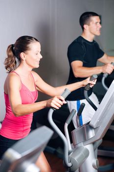 People - man and woman - exercising in a gym on an elliptical trainer for more fitness