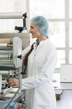Happy female manufacturing operator wearing white lab coat and protective headwear, while working with modern equipment in a cosmetics factory