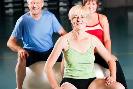 Three people - mature and senior - in the gym doing gymnastics on an exercise ball