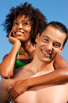 Couple in love - bikini-clad woman of color hugs a Caucasian man from behind under clear blue sky, both in beachwear in summer FOCUS ON MAN